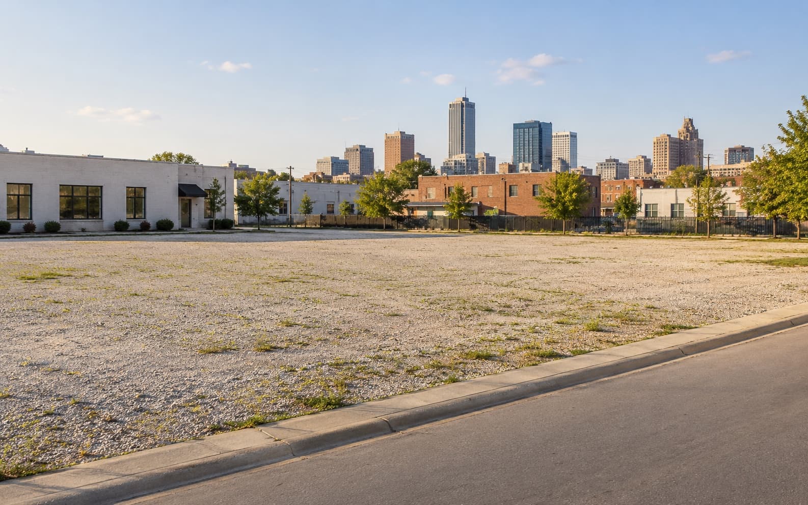 An open gravel lot with low commercial buildings and a city skyline behind it.